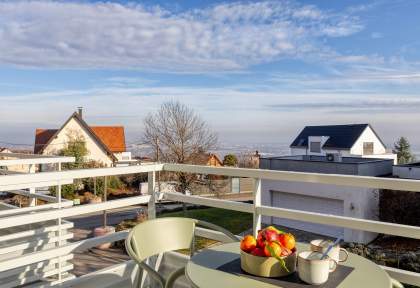 Balcon d’un appart-hôtel avec table, chaises, corbeille de fruits et vue dégagée sur les environs résidentiels de la pleine d'Alsace