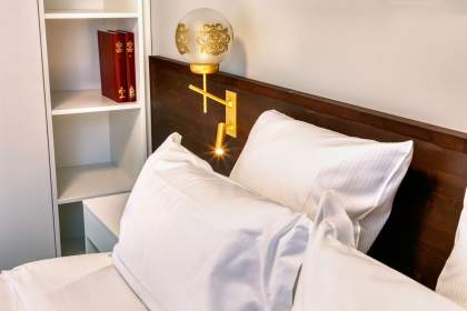 Détail de la chambre d’un appart-hôtel avec tête de lit, lampe d’ambiance, linge blanc et étagère décorative près de Colmar, en Alsace