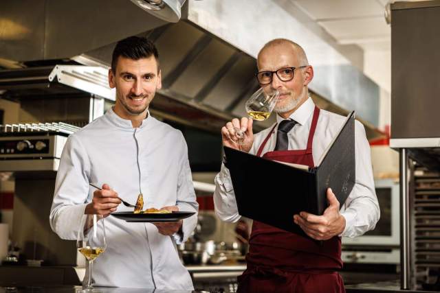 Chefkoch und Sommelier in einer Profiküche präsentieren ein Gericht und ein Glas Wein – ein kulinarisches Erlebnis in der Nähe von Colmar im Elsass.