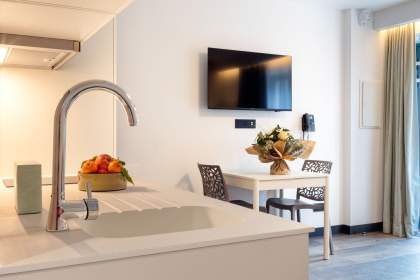 Coin repas d’un appart-hôtel avec table, chaises design, télévision murale et cuisine équipée à proximité près de Colmar, en Alsace