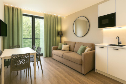 Séjour lumineux d’un appart-hôtel avec canapé confortable, coin repas, baie vitrée et vue sur la verdure près de Colmar, en Alsace